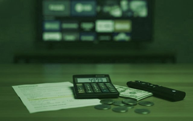 A calculator, dollar bills, coins, a streaming stick and a remote on a table in front of a TV